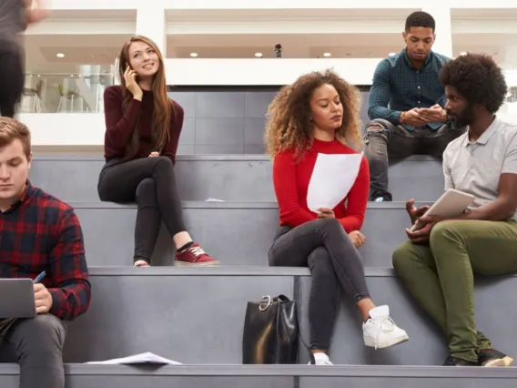 A diverse group of students are sitting on the steps of a conference room, while discussing and reviewing their projects.