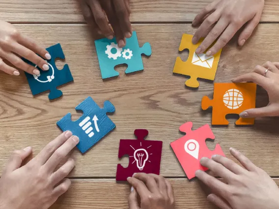 A group of six hands are holding colored puzzle pieces with different iconography on a wooden table.