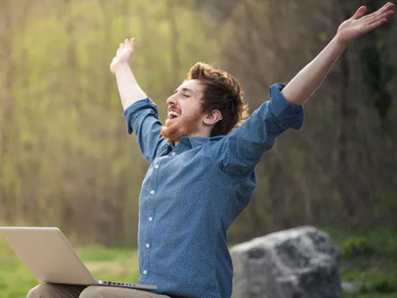 A student that is working outdoors on his laptop raises his arms and smiles happily and freely. 