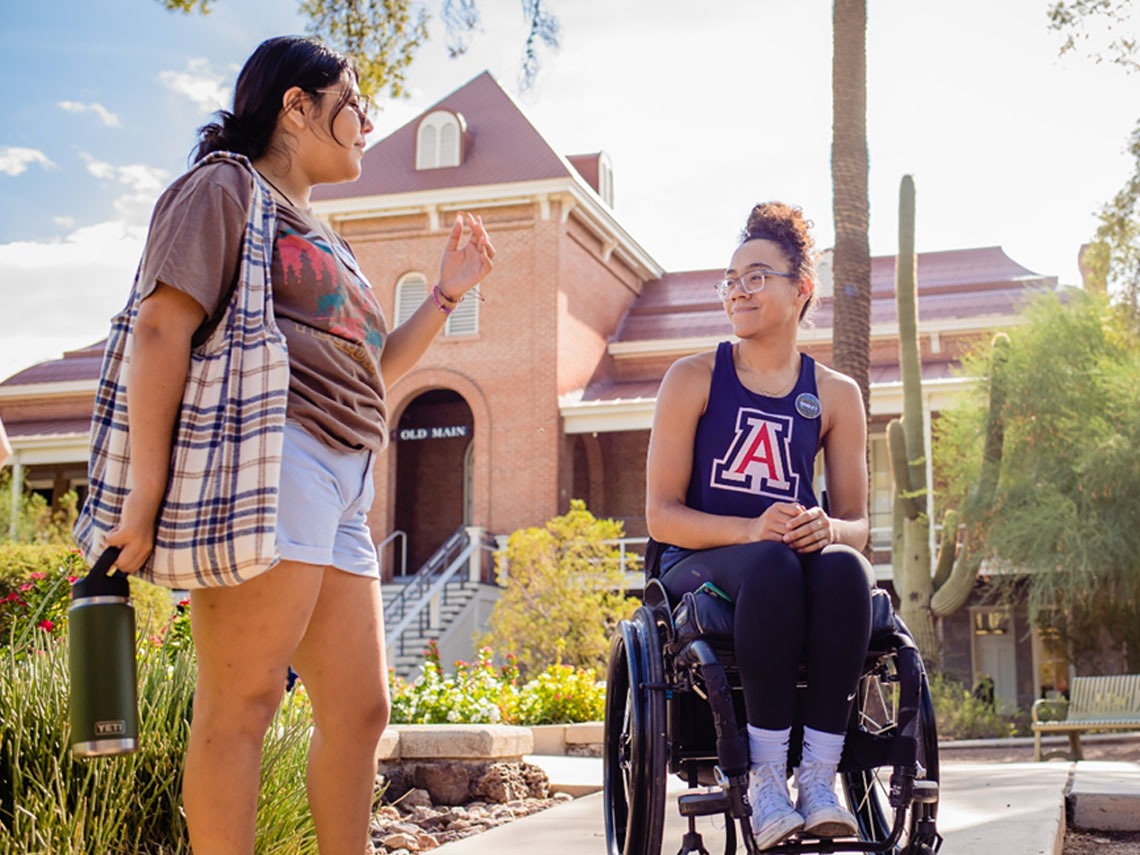 A student in a wheelchair talks to her classmate about their college experiences by the water fountain in front of Old Main.