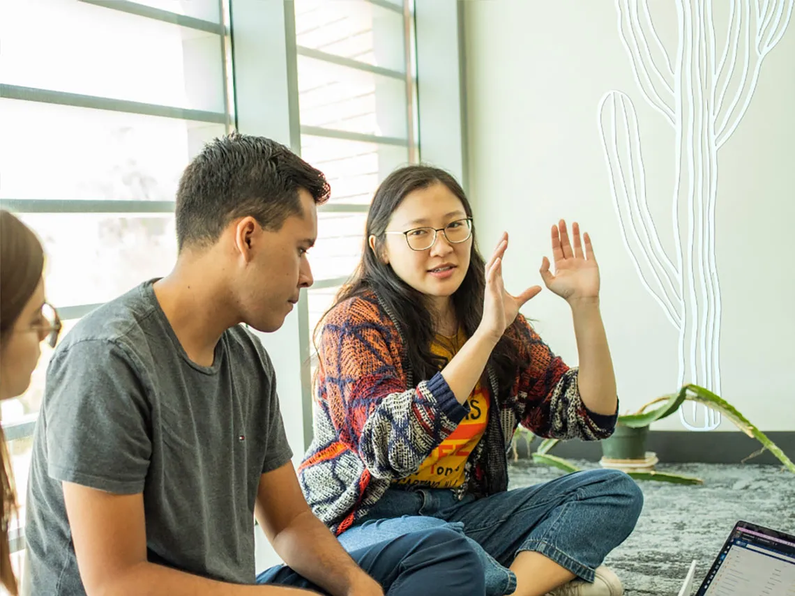 A discussion about an assignment is taking place among a few students. They are sitting on the floor of a room while they review their task on their laptop computer.