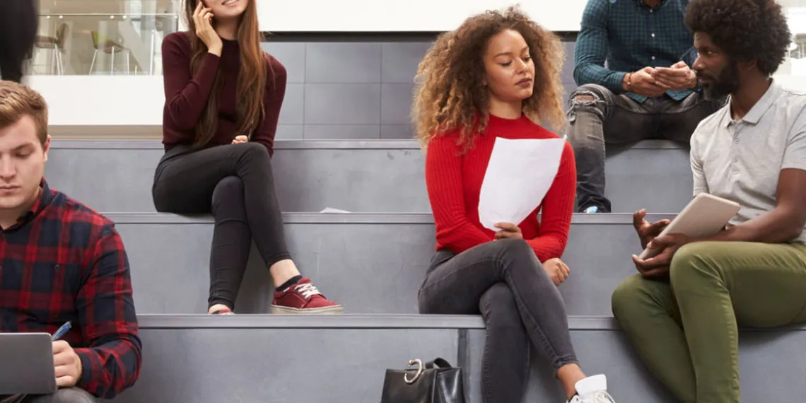 A diverse group of students are sitting on the steps of a conference room, while discussing and reviewing their projects.
