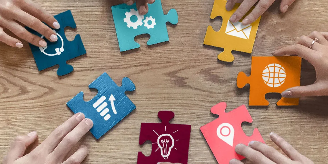 A group of six hands are holding colored puzzle pieces with different iconography on a wooden table.