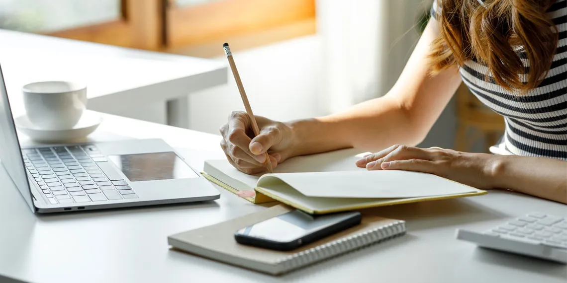 A faculty is taking notes on a notepad while prepping her online class session.