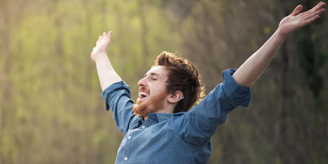 A student that is working outdoors on his laptop raises his arms and smiles happily and freely. 